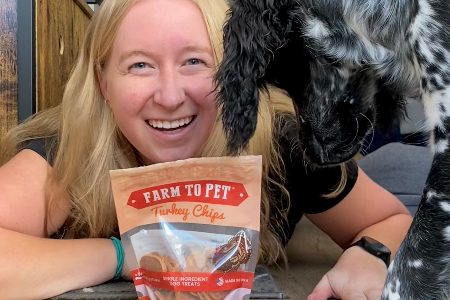 Blonde woman smiling while giving her black spotted dog Farm to Pet turkey chips