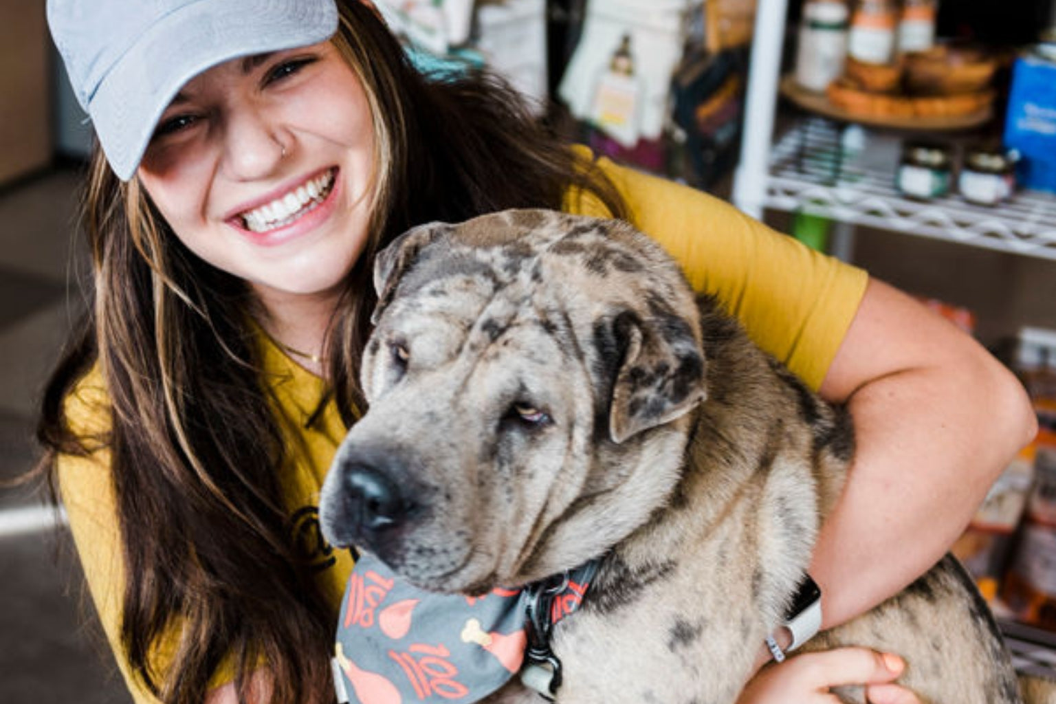 A smiling woman in a hat hugging her dog in a Farm to Pet bandana.